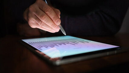 Male hand holds stylus over screen of tablet with commercial charts in dark. Deadlines at work...