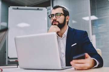 Thoughtful businessman dressed in suit thinking on productive strategy for developing own financial company working at modern laptop computer connected to wireless 4G internet in stylish office