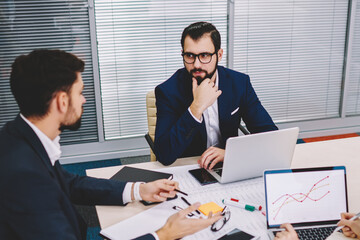 Owner of financial company talking with business partners about developing new project during brainstorming in office sitting at meeting table with laptops.Proud ceo discussing plan with colleagues