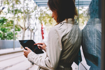 Fototapeta premium Stylish young woman dressed in coat making payment online on modern touch pad device via 4G internet standing in urban setting on tram stop.Hipster girl chattin online on digital tablet on street