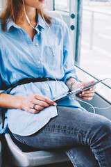 Cropped view of young woman passenger in earphones listening audio book downloaded on modern tablet sitting in tram.Hipster girl holding touch pad in hands updating profile via public transport wifi