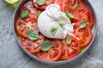 Caprese with sliced burrata cheese, red tomatoes and fresh green basil, middle close-up, horizontal shot on a grey granite surface