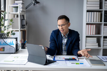 A man in a suit is sitting at a desk with a laptop and papers