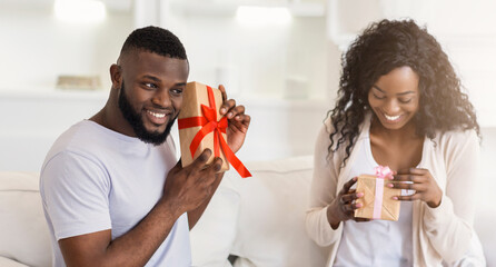 A man smiles and presents a gift to a woman who is opening it with excitement.