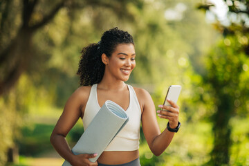 A young woman, dressed in athletic wear, holds a yoga mat and checks her phone in a lush park setting. She appears to be checking her schedule or a yoga app before starting her session.