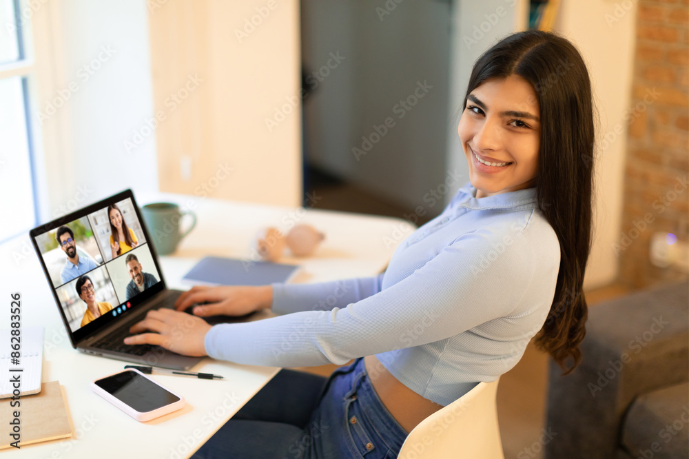 Poster smiling woman sits at a desk in a home office, smiling and looking at the camera while participating