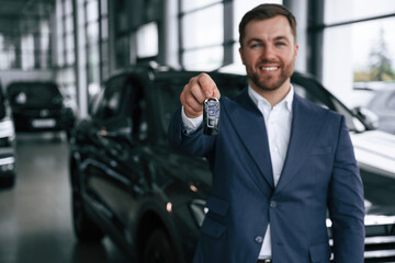 Owning the automobile, holding keys. A businessman is in a car dealership