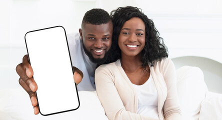 A smiling Black couple holds a smartphone with a blank screen. The couple is sitting on a white...