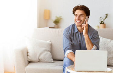 A man sits on a couch in a living room, talking on his phone while using a laptop.
