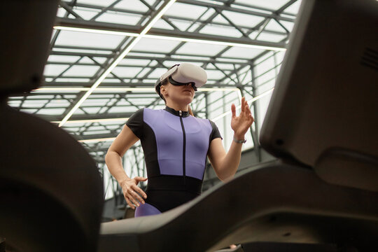 Graphic low angle portrait of sportive woman wearing VR headset and running on treadmill in futuristic gym against glass ceiling
