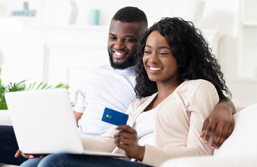 Obraz premium A couple sits on a couch, smiling, as they use a laptop to shop online. The woman is holding a credit card.