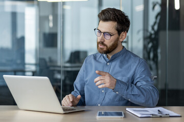 Serious and focused young businessman in glasses and blue shirt sitting in the office at the table and having a video call, conference and online meeting