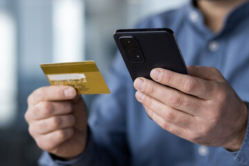 A close-up photo of a body part. Hands of a young businessman in a blue shirt holding a credit card and using a mobile phone