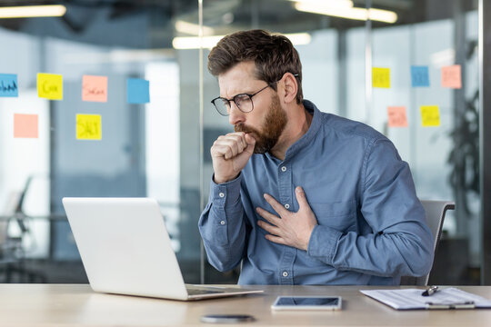 A young male office worker sits at a desk at work and coughs, covers his mouth with his hand and holds his chest, breathing hard and feeling unwell