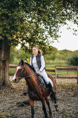 A young beautiful woman jockey is preparing for a show jumping competition. A woman rider rides a brown racehorse.