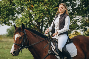 Young beautiful blonde woman jockey rides a brown horse in a meadow at sunset in summer. Preparing...