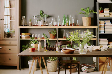 Wooden table with perfumery items and testers, shelves full of bottles with essential oil and bookcase with books and notebooks in home perfume lab