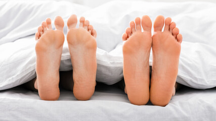 This image shows two pairs of bare feet sticking out from under white bed sheets. The feet are positioned close together, suggesting that the couple is in bed together.