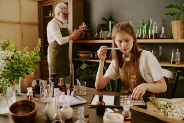 Female perfumer sitting at table and smelling tester while mature male perfumer reading notes