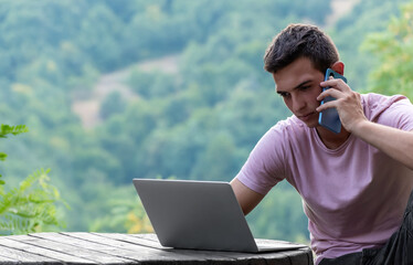 Young man work on laptop and talk on phone sitting at wooden table in mountains nature outside....