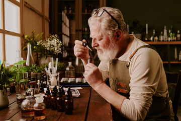 Senior perfumer sitting at wooden table with plants and bottles with samples and smelling new scent while glass pipette trickling down aromatic liquid