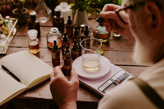Over shoulder shot of mature perfumer adding extra essential oil while glass flask with perfume sample on kitchen scales according to his notes
