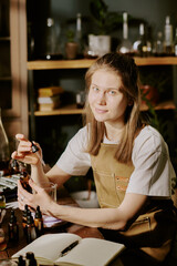 Young female perfumer sitting at table and holding brown glass bottle of perfume sample, she smiling gently and enjoying process