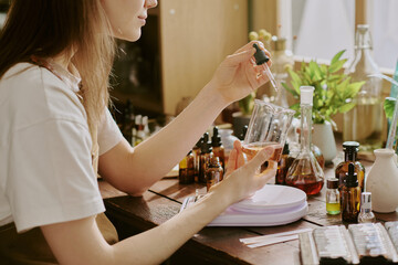 Young female perfumer sitting at table and holding glass pipette with essence and adding it into caramel aromatic essence