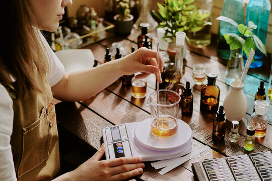 Young female perfumer blending essential oils while she weighing it with plastic kitchen scale
