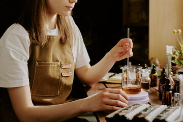Female perfumer sitting at table and mixing caramel shade essence, she surrounded by perfume...