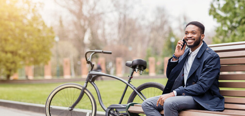 Obraz premium An African American businessman is talking on his phone while sitting on a bench in a park, with his bicycle nearby. The mood is relaxed and professional, set in a green, peaceful park.