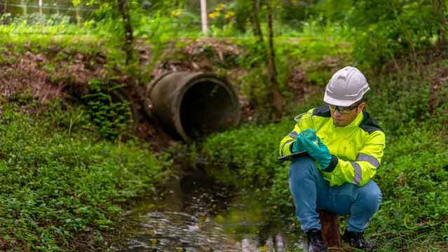 An ecologist in a green jacket sitting on a steel pole while holding the plastic glass on his left hand examination the quality of the water. and write down the result on a clipboard