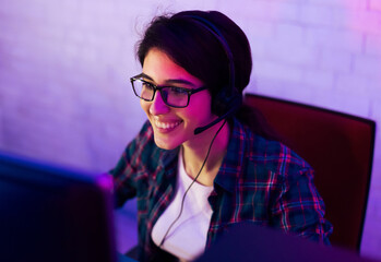 A young woman with dark hair is wearing glasses and a headset while sitting in front of a computer. She is smiling and looking at the screen. The room is lit in a purple and pink glow
