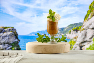 Glass of iced coffee with cream and mint leaves on seascape mountains island background and copy space for products.