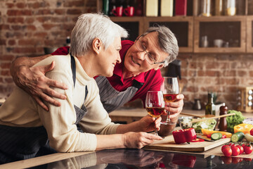 A senior couple is enjoying a glass of wine together in their kitchen. The man is leaning in towards his wife and smiling as they both raise their glasses.
