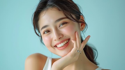 Japanese woman in her late thirties, smiling and pointing to the side of her face with one finger while looking at camera. She is wearing a white sleeveless top on a light blue background. The focus