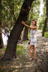 An attractive young woman in a summer floral dress happily stands by a tree on a serene nature trail, exuding joy and tranquility.