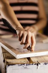 Close-up of a female painter's hand delicately using a palette knife on a canvas propped in her art studio, surrounded by various painting supplies.