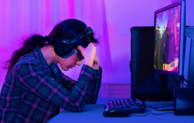 A young woman sits at a computer desk with her head in her hands, looking defeated. She is wearing headphones and glasses and is likely playing a video game on her computer.