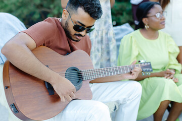 Multiracial group of people, indian man playing guitar and friends eating dinner and drinking wine during party in the picnic