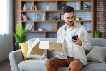 Obraz premium Angry young Indian man sitting on sofa at home, holding open parcel box and looking disappointedly at mobile phone, dialing delivery service with complaints