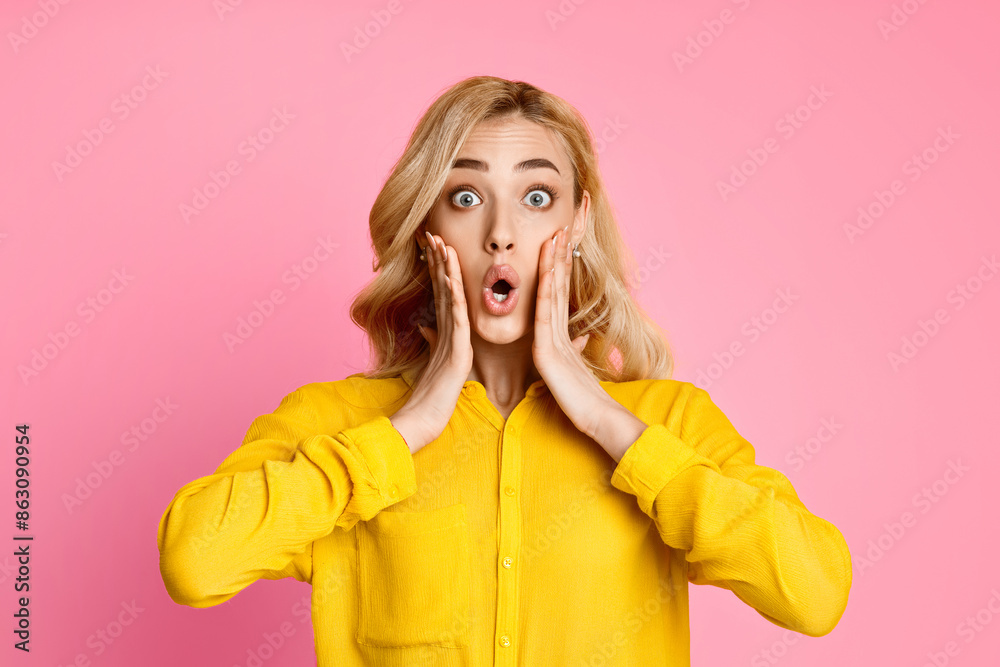 Wall mural This image shows a woman in a yellow shirt with a surprised expression on her face. Her hands are placed on her cheeks, and her eyes are wide open. The background is a solid pink.