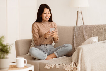 A young woman is sitting on a couch in a living room and using a phone. She is smiling and looking...