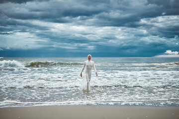 Hairless girl with alopecia in white futuristic suit comes out of cold restless sea on sandy beach,...