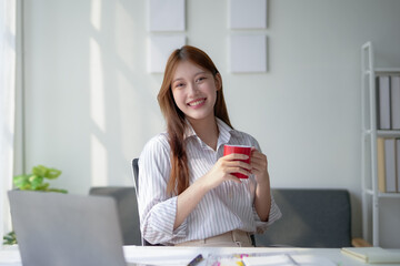 Smiling woman holding a red mug, sitting at a desk with a laptop in a bright office setting. Ideal...