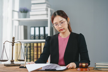 Young female lawyer working at desk with legal books, scales of justice, and gavel, focused on paperwork in modern office.