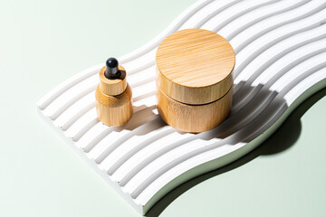 Cosmetic set of face beauty oil in bamboo dropper bottle and face cream in a jar on white curved concrete tray on green background, Unbranded skin care treatment packaging