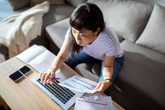 Concerned Asian woman holding bills and using laptop at home