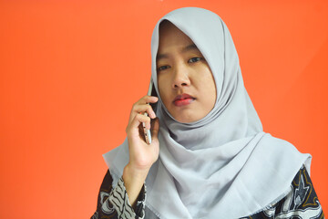 Dissatisfied young Asian girl standing holding mobile phone. Isolated on orange background