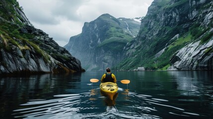 Kayaker paddling a yellow kayak on a calm fjord, with steep, green, cliff-like mountains on either side, waterfalls, and a cloudy sky